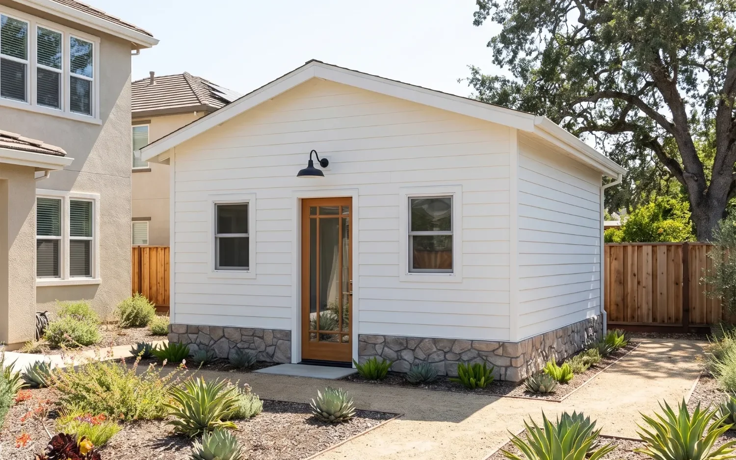 A finished white detached ADU with a stone base and a wood-framed glass door, beside a mature oak