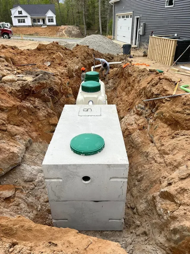 A two-piece precast concrete septic tank set at the bottom of a deep trench, its lid fitted with a green access riser, with a plastic tank and green risers behind it and a crew connecting the PVC line