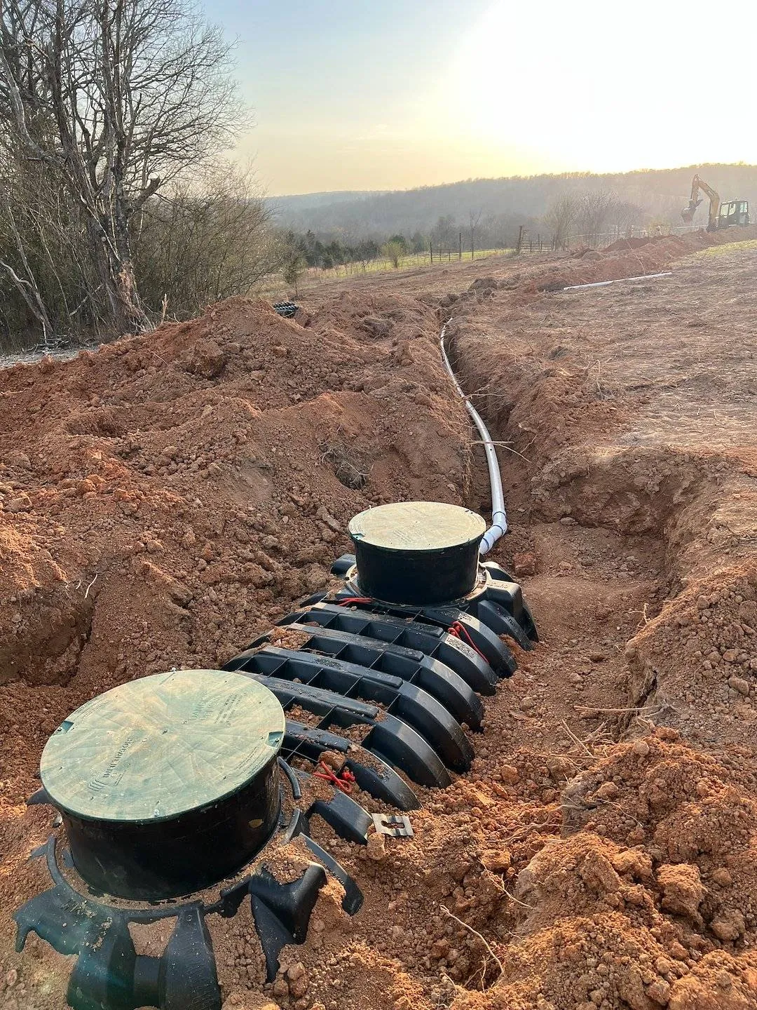 A new septic tank with two green access lids set in a freshly dug trench of red clay, its white drain line running toward an excavator in the distance
